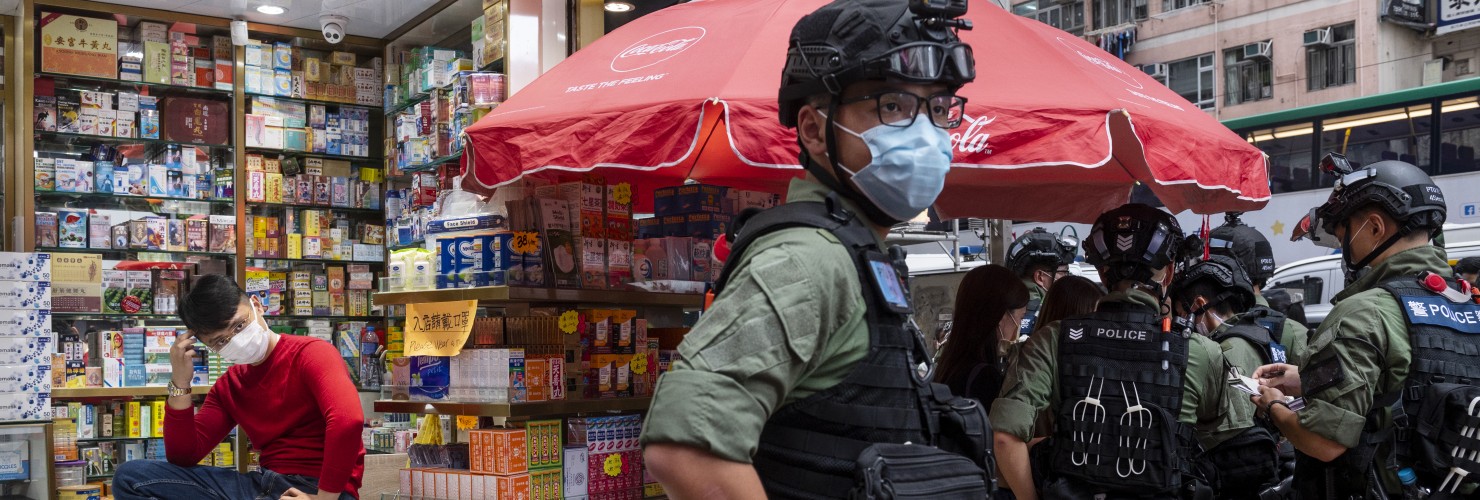 A pharmacy sales person sits on a chair as riot police officers stop and search people on the vicinity during a banned protest in Hong Kong, China, on October 1, 2020.